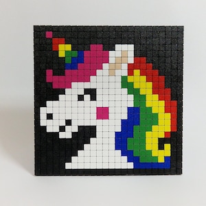 Könnte beinhalten: Ein pixeliges Einhorn-Design auf schwarzem Hintergrund. Das Einhorn ist weiß mit einer rosa Mähne, einem Regenbogenhorn und einem rosa Rouge. Das Kunstwerk besteht aus kleinen, quadratischen Kacheln.