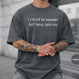 Może przedstawiać: Ciemnoszara koszulka z krótkim rękawem z białym napisem: "I could be meaner but Jesus said no". Koszulka ma luźny krój i efekt vintage. Kolor koszulki to pieprz.