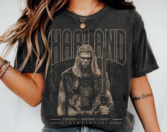May include: A charcoal grey short-sleeve t-shirt featuring a graphic print of a long-haired warrior holding a sword. The text reads "HAALAND" at the top and "STRENGTH HERITAGE LEGACY VIKING SPIRIT" at the bottom in a distressed, vintage-style font.
