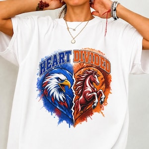 以下が含まれることがあります： 2つに分かれたハートのグラフィックが描かれた白いTシャツ。左側は青色でハクトウワシ、右側はオレンジ色で後ろ足で立つ馬が描かれています。ハートのデザインの上には「HEART DIVIDED」という文字が太字でプリントされています。