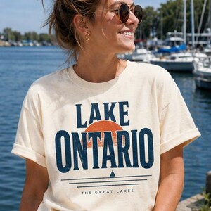 May include: A cream-colored short-sleeve t-shirt featuring the text LAKE ONTARIO in dark blue, a semi-circular orange sun graphic, a small sailboat icon, and the text THE GREAT LAKES printed in a smaller font below the horizontal lines.