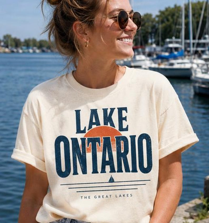 May include: A cream-colored short-sleeve t-shirt featuring the text LAKE ONTARIO in dark blue, a semi-circular orange sun graphic, a small sailboat icon, and the text THE GREAT LAKES printed in a smaller font below the horizontal lines.