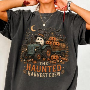 Puede incluir: Una camiseta gris carbón con un gráfico de un fantasma con sombrero de bruja conduciendo un tractor que tira de un remolque lleno de calabazas. El diseño incluye un gato negro, murciélagos, una luna creciente y el texto THE HAUNTED HARVEST CREW.