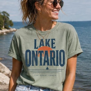 May include: A sage green short-sleeve t-shirt featuring the text LAKE ONTARIO in dark blue block letters. A graphic of an orange sun and a small sailboat sits between the words. The text THE GREAT LAKES appears in smaller font below.