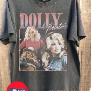 Puede incluir: Una camiseta de manga corta en color gris carbón con un estampado gráfico de Dolly Parton. El diseño incluye el texto "DOLLY Parton" sobre tres imágenes de la artista. La camiseta tiene cuello redondo y corte recto, con un logotipo "MADE IN USA".