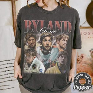 May include: Dark gray t-shirt featuring a graphic design with the text "RYLAND Grace" in a vintage style. The design includes multiple images of a person. The shirt has a crew neck and short sleeves.
