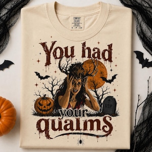 May include: A beige t-shirt featuring a graphic print with the text "You had your qualms" in a dark brown, dripping font. The illustration depicts a person with antlers, a large orange moon, bare trees, bats, a jack-o'-lantern, and a tombstone.