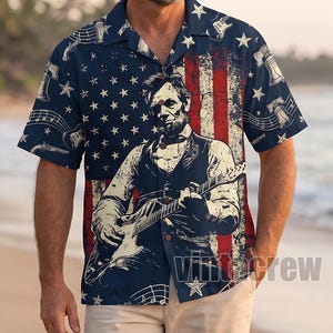 May include: Navy blue short-sleeve shirt with a patriotic design. The shirt features a distressed American flag, stars, musical notes, and a graphic of Abraham Lincoln playing a guitar. The shirt has a button-down collar and is worn by a person on a beach.