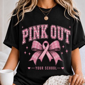 Op de afbeelding: Een zwart t-shirt met korte mouwen en de tekst PINK OUT in een roze en wit ruitpatroon. Onder de tekst staat een grote roze strik met een ruitpatroon. De tekst YOUR SCHOOL staat onderaan met kleine hartjes.