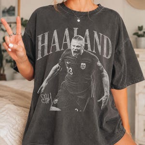 May include: A charcoal gray oversized t-shirt featuring a distressed white graphic print of a soccer player in a jersey with the number 23. The text HAALAND is printed in a large arched font above the player, with a signature graphic below.