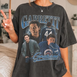 Puede incluir: Una camiseta gris carbón de corte oversize con un estampado gráfico de estilo vintage que muestra el nombre GARRETT en letras azules en la parte superior. El diseño incluye varias imágenes fotográficas de un hombre y el nombre Graham en azul.