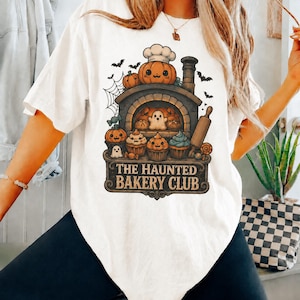 May include: A white t-shirt featuring a graphic of a stone oven with a pumpkin chef wearing a hat on top. The oven contains a ghost and baked goods, with cupcakes, candy, and bats surrounding it. The text reads THE HAUNTED BAKERY CLUB.