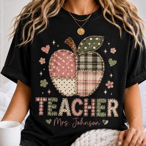 May include: A black t-shirt featuring a large graphic of an apple made of various patterned patches including polka dots and plaid. Below the apple, the word TEACHER is printed in a patchwork style, followed by the text Mrs. Johnson in script.