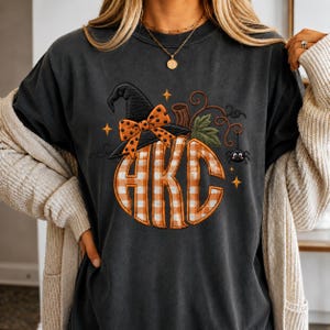 May include: A charcoal gray t-shirt featuring an embroidered pumpkin graphic with a white and orange gingham pattern. The pumpkin is topped with a black witch hat adorned with an orange polka dot bow, a green leaf, and a small black spider.