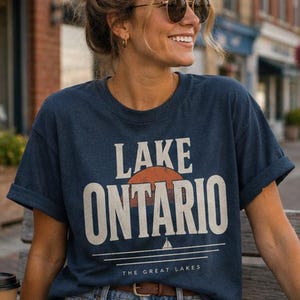 May include: A navy blue short-sleeved t-shirt featuring a graphic print that reads LAKE ONTARIO in large white letters. The design includes a burnt orange sun, a small white sailboat icon, and the text THE GREAT LAKES at the bottom.