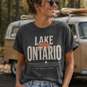 May include: A dark charcoal gray short-sleeve t-shirt featuring a graphic print with the text LAKE ONTARIO in large white letters. Below the text is a stylized orange sun, a small white sailboat icon, and the text THE GREAT LAKES.