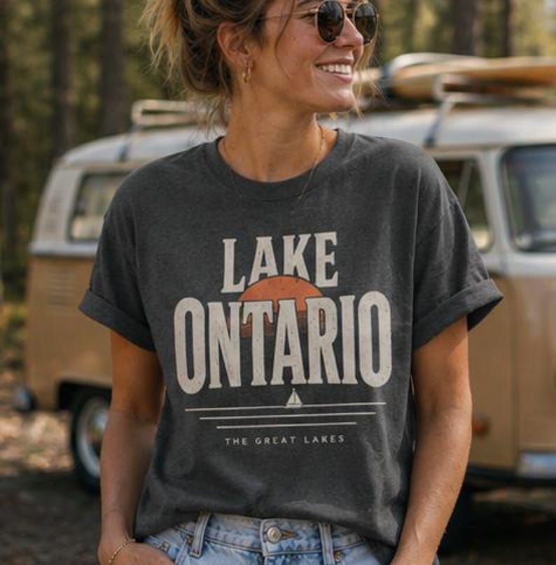 May include: A dark charcoal gray short-sleeve t-shirt featuring a graphic print with the text LAKE ONTARIO in large white letters. Below the text is a stylized orange sun, a small white sailboat icon, and the text THE GREAT LAKES.