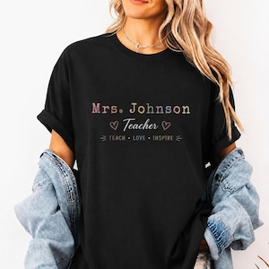 Może przedstawiać: Czarny t-shirt z krótkim rękawem z napisem Mrs. Johnson wielokolorowymi literami, słowem Teacher białym pismem odręcznym oraz słowami TEACH LOVE INSPIRE białymi wielkimi literami, ozdobiony dwoma małymi białymi konturami serc.