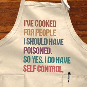 May include: Cream-colored apron with the text "I'VE COOKED FOR PEOPLE I SHOULD HAVE POISONED. SO YES, I DO HAVE SELF CONTROL." A pen is in the pocket. The apron is on a wooden surface with cooking utensils and food.