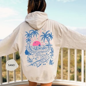 May include: A sand-colored hoodie with a beach scene graphic on the back. The design features palm trees, a sunset, a sailboat, and a winding path. The word "SAND" is printed on the front.