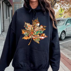 May include: A black hooded sweatshirt featuring a large, central graphic of a maple leaf. The leaf is filled with a patchwork pattern of autumn-themed designs, including pumpkins, plaid, and floral motifs. The hoodie includes a front kangaroo pocket.