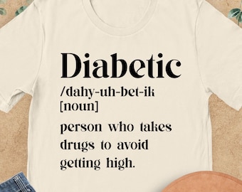 May include: A cream-colored t-shirt with the word "Diabetic" in bold black letters, along with a definition. The shirt is paired with blue jeans and a brown hat. The text on the shirt reads: "person who takes drugs to avoid getting high."