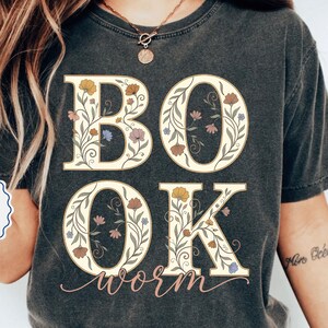 以下が含まれることがあります： 大きな花柄の文字で「BOOK」と「OK」と書かれた濃い灰色のTシャツ。「worm」の文字が筆記体で下に書かれています。シャツはヴィンテージ風で、色あせた外観です。「MORE COLOR OPTIONS」の文字が見えます。