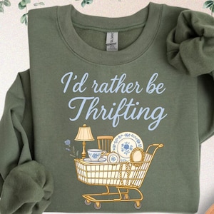 May include: Military green sweatshirt with the text "I'd rather be Thrifting" in light blue script. A shopping cart graphic is filled with vintage items like a lamp, dishes, and a teacup. The sweatshirt is folded, with a pair of jeans and a hat in the background.