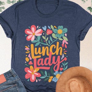 Pode incluir: T-shirt azul marinho com design floral e a frase "Lunch Lady". A imagem tamb&eacute;m inclui um par de jeans azuis e um chap&eacute;u castanho. A t-shirt &eacute; feita de um material macio.