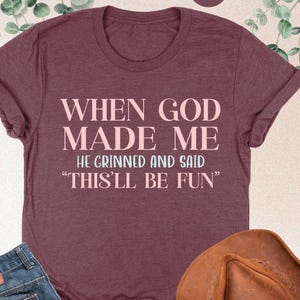 May include: Heather maroon t-shirt with the text "WHEN GOD MADE ME HE GRINNED AND SAID 'THIS'LL BE FUN'". The image also includes a pair of blue jeans and a brown cowboy hat.