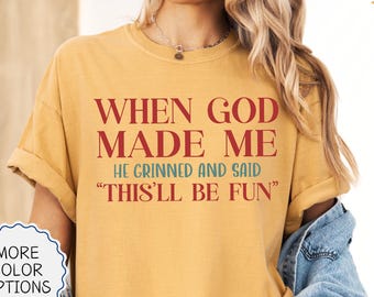 May include: Mustard-colored t-shirt with the text "WHEN GOD MADE ME HE GRINNED AND SAID 'THIS'LL BE FUN'" in red. The shirt has rolled-up sleeves and is part of a clothing collection. Additional color options are available.