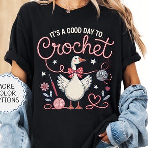 May include: Black t-shirt with the text "IT'S A GOOD DAY TO. Crochet" in a whimsical font. The graphic features a goose wearing a bow tie, surrounded by yarn balls, flowers, and crochet hooks. The words "MORE COLOR OPTIONS" are also visible.