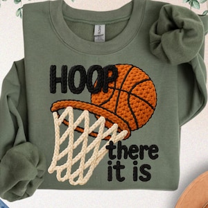 May include: Military green sweatshirt with a basketball hoop design. The design features a basketball and net with the words "HOOP there it is" in black. The sweatshirt is made of faux yarn.
