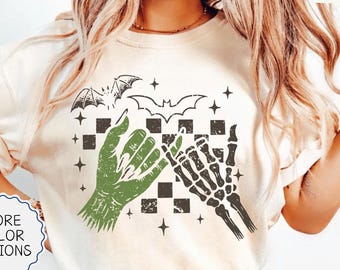 May include: An ivory t-shirt featuring a graphic print of a green hand and a skeletal hand performing a pinky promise. The design includes a black and white checkered pattern, two bats, and several small stars. The text IVORY appears in the corner.