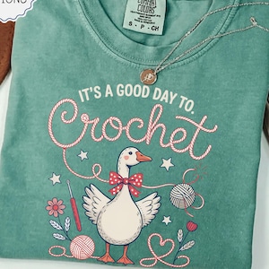 May include: Light green t-shirt with the phrase "IT'S A GOOD DAY TO. Crochet" and a cartoon goose wearing a bow tie. The shirt also features yarn balls, crochet hook, and stars. A silver necklace is draped across the shirt.