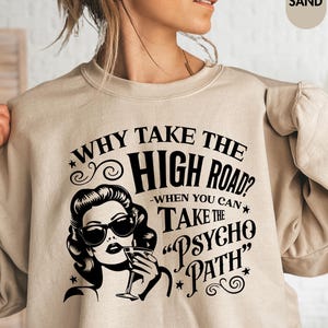 May include: Sand-colored sweatshirt with the text "WHY TAKE THE HIGH ROAD? WHEN YOU CAN TAKE THE PSYCHO PATH" and a black and white graphic of a woman wearing sunglasses and holding a drink.