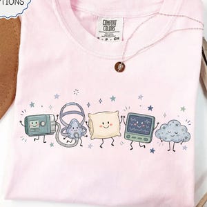 May include: A light pink crew neck t-shirt featuring a graphic print of five cartoon characters: a CPAP machine, a nasal mask, a pillow, a heart rate monitor, and a cloud, all with smiling faces, surrounded by small blue stars and sparkles.