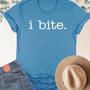 May include: A heather deep teal t-shirt with the text "i bite." is displayed with a pair of blue jeans and a tan fedora hat. The shirt is folded at the sleeves. The jeans have a red leather patch.