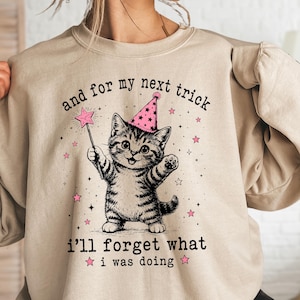 May include: A sand-colored crewneck sweatshirt featuring a graphic of a tabby kitten wearing a pink party hat, holding a star-tipped magic wand. The text reads, and for my next trick i'll forget what i was doing, surrounded by small pink stars.