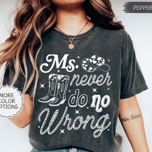 May include: Dark gray t-shirt with the text "Ms. never do no Wrong" in white. The design includes cowboy boots, a cowboy hat, and stars. The shirt has a vintage, worn look.