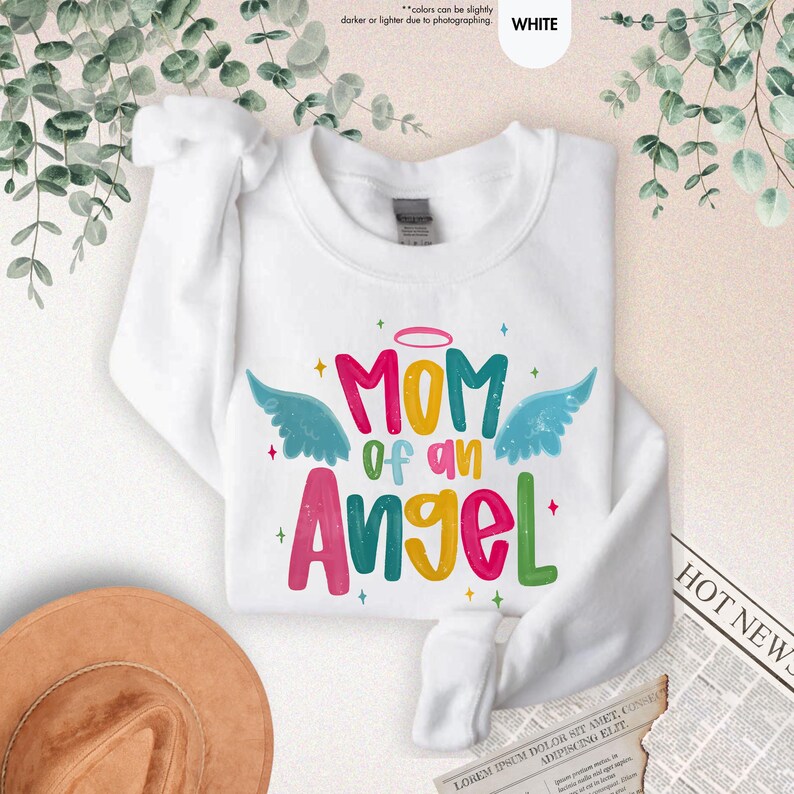 May include: White sweatshirt with the text "MOM of an Angel" in colorful letters, with angel wings and a halo design. The sweatshirt is folded and displayed on a light background.