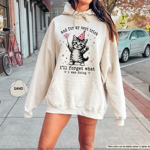 May include: A sand-colored hooded sweatshirt featuring a graphic of a tabby cat wearing a pink party hat and holding a magic wand. The text reads, and for my next trick i'll forget what i was doing, surrounded by small pink stars.