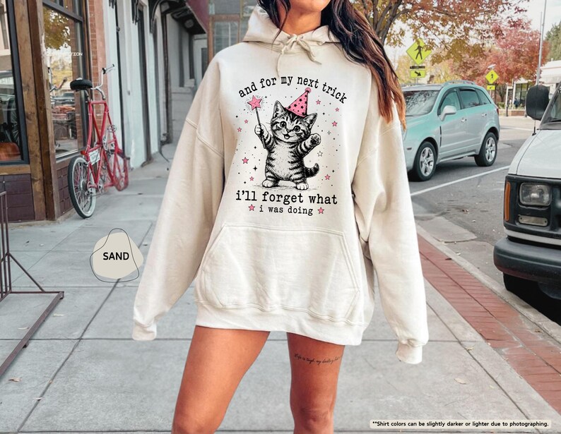 May include: A sand-colored hooded sweatshirt featuring a graphic of a tabby cat wearing a pink party hat and holding a magic wand. The text reads, and for my next trick i'll forget what i was doing, surrounded by small pink stars.
