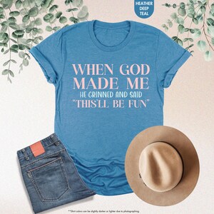 May include: A heather deep teal t-shirt with the text "WHEN GOD MADE ME HE GRINNED AND SAID 'THIS'LL BE FUN'" in pink. The shirt is paired with blue jeans and a tan hat. The shirt's color may vary slightly.