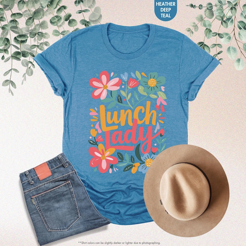 Pode incluir: Uma t-shirt azul-petr&oacute;leo com a frase "Lunch Lady" rodeada por flores coloridas. A imagem tamb&eacute;m inclui um par de cal&ccedil;as de ganga azuis e um chap&eacute;u bege.