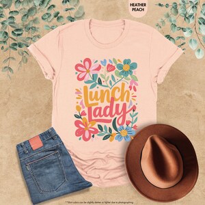 Pode incluir: Camiseta cor de p&ecirc;ssego com um design floral e a frase "Lunch Lady" em uma fonte colorida. A camiseta &eacute; combinada com jeans azuis e um chap&eacute;u marrom. A camiseta &eacute; feita de um material macio.