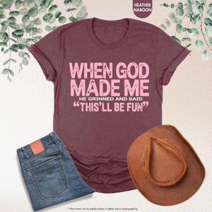 May include: A maroon t-shirt with the text "WHEN GOD MADE ME HE GRINNED AND SAID 'THIS'LL BE FUN'" in pink. The image also includes a pair of blue jeans and a brown cowboy hat.