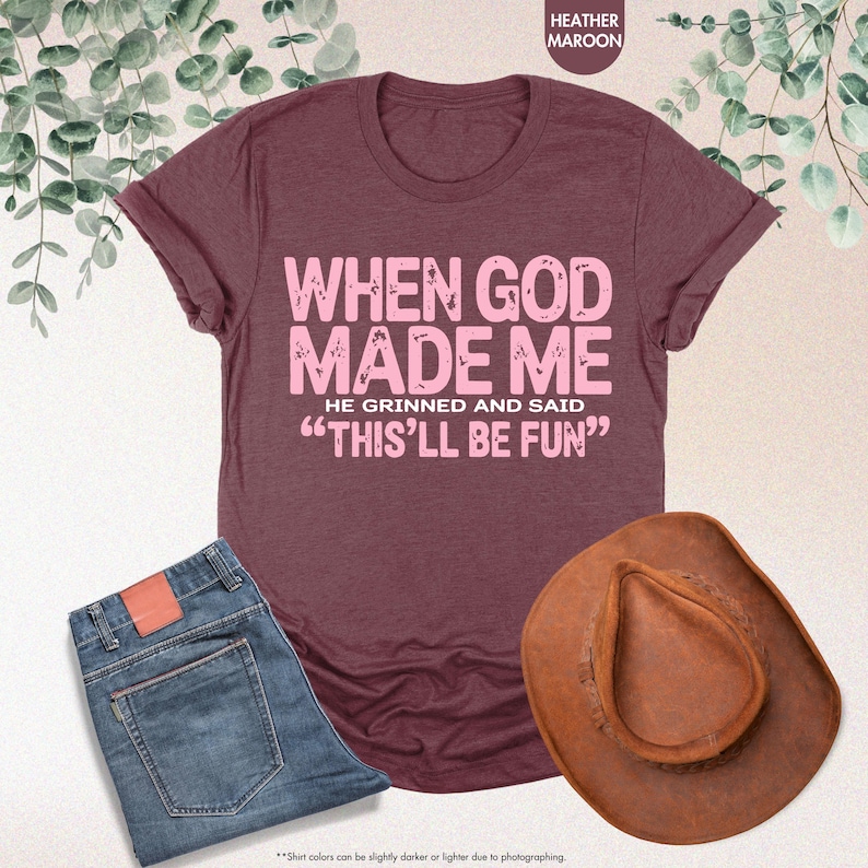 May include: A maroon t-shirt with the text "WHEN GOD MADE ME HE GRINNED AND SAID 'THIS'LL BE FUN'" in pink. The image also includes a pair of blue jeans and a brown cowboy hat.