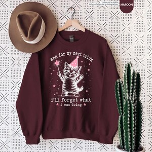 May include: A maroon crewneck sweatshirt featuring a white graphic of a kitten wearing a pink party hat and holding a star wand. The text reads, and for my next trick i'll forget what i was doing, surrounded by small pink stars.