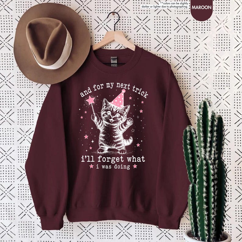 May include: A maroon crewneck sweatshirt featuring a white graphic of a kitten wearing a pink party hat and holding a star wand. The text reads, and for my next trick i'll forget what i was doing, surrounded by small pink stars.
